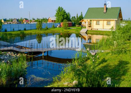 Un chalet en bois avec un belvédère se trouve dans un jardin près d'un étang avec un pont et des fleurs au printemps. Banque D'Images