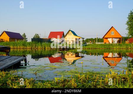 Un petit quai fait de planches dans le jardin à côté de l'étang au milieu de l'herbe et des fleurs de nénuphars sur la toile de fond de chalets en bois. Banque D'Images