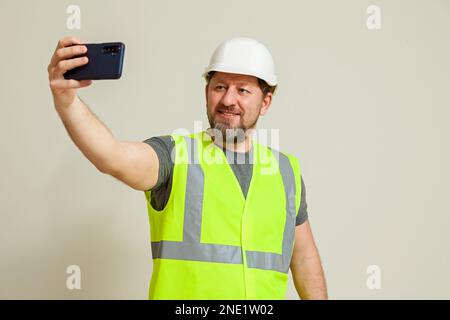 Un homme travailleur dans un gilet et un casque de construction blanc prend un selfie sur le téléphone sur un fond blanc Banque D'Images