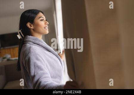 Vue latérale de la jeune femme du Moyen-Orient regardant par la fenêtre à la maison. sans modification, style de vie, contemplation, rêve de jour, espace de copie. Banque D'Images