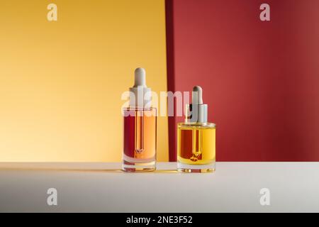 Huile pour le visage et les cheveux rose et jaune dans des flacons en verre avec pipette compte-gouttes sur fond orange vif et rouge. Photo d'auto-soin de beauté basse clé Banque D'Images