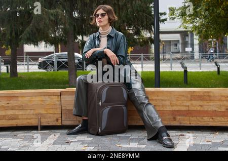 Une femme en cuir et avec une valise est assise sur un banc de parc Banque D'Images