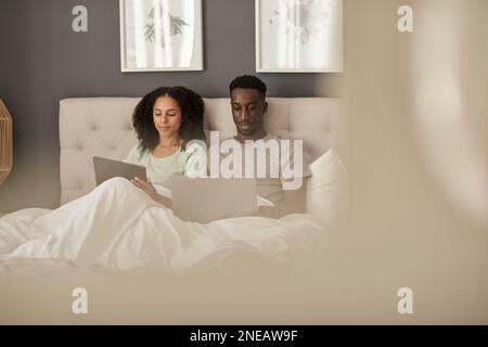 Couple multiracial travaillant sur différents appareils au lit le matin Banque D'Images