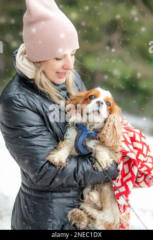 Portrait d'une femme d'âge moyen souriante vêtue d'une veste noire, tenant en forêt un hiver enneigé pour observer l'épagneul de chien. Banque D'Images