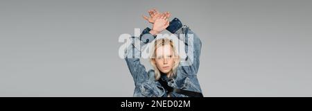 jeune femme blonde dans une élégante veste en denim regardant l'appareil photo tout en posant avec les mains au-dessus de la tête isolée sur gris, bannière, image de stock Banque D'Images