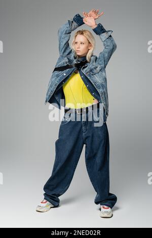 pleine longueur de femme blonde dans une tenue élégante en denim posant les mains au-dessus de la tête sur une image grise Banque D'Images