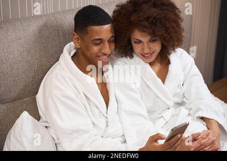 Un adorable couple multiracia dans des peignoirs est assis sur le lit Banque D'Images