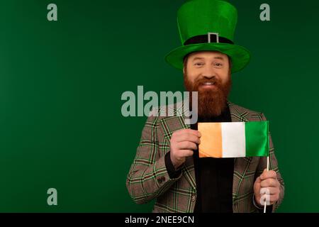 Homme positif dans le chapeau tenant le drapeau irlandais pendant la fête de saint patrick isolé sur vert, image de stock Banque D'Images