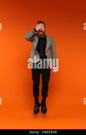 Homme barbu excité couvrant les yeux tout en sautant sur fond rouge, image de stock Banque D'Images