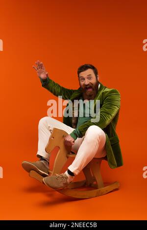 Homme barbu excité dans la veste en agitant la main tout en étant assis sur cheval de bascule sur fond rouge, image de stock Banque D'Images