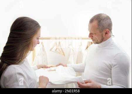 magasin de vêtements jeune fille vendeur offre un t-shirt blanc à un homme homme frowning examine sérieusement ses rides de barbe sur son visage sensibilité d'une jeune fille à un homme 50-60 ans filles 20-25 studio Banque D'Images