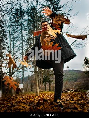 Une photo verticale d'un jeune homme qui lève des feuilles d'automne sèches dans une forêt Banque D'Images