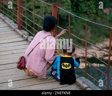 Un garçon habillé comme batman et sa grand-mère regardent sur une rivière depuis un pont à Muang Khua, au Laos. Banque D'Images
