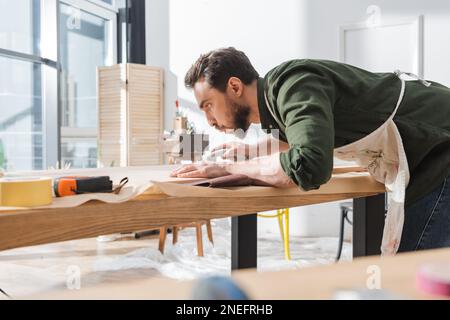 Vue latérale du restaurateur barbu soufflant de la poussière du panneau en bois près du papier de verre dans l'atelier, image de stock Banque D'Images