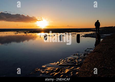 Ciel orange au-dessus de Worthing, West Sussex en hiver avec un homme en silhouette marchant le long de la plage Banque D'Images