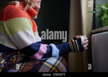 Femme en gants avec boisson chaude essayant de rester au chaud par le radiateur pendant la crise énergétique du coût de la vie Banque D'Images