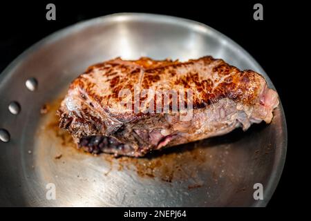 Gros plan d'une poêle en acier inoxydable et d'une croûte sur une épaisse bande de New york ou un steak de viande de ribeye cuisant sur une cuisinière avec texture charrée et fond noir Banque D'Images