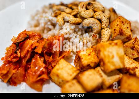 Cubes de tofu faits maison, noix de cajou, riz brun, chou kimchi mariné sur plaque blanche macro gros plan avec texture détail de la nourriture Banque D'Images