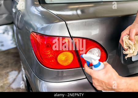 Homme lavant le véhicule à la station de lavage automatique, vaporisant la solution de nettoyage en mousse du flacon de pulvérisation sur les feux arrière du coffre à l'extérieur, essuyant Banque D'Images