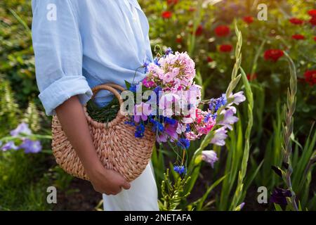Gros plan d'un sac de paille rempli de fleurs bleues et roses. Femme tenant sac à main d'été avec bouquet de fleurs. Accessoires tendance pour femme Banque D'Images