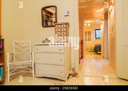 Chaise en osier peint en blanc à côté d'une commode en bois antique dans la chambre principale et vue sur la chambre d'amis et le couloir avec de larges planchers peints. Banque D'Images