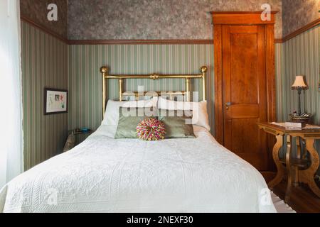 Lit double avec couvre-lit blanc et tête de lit en laiton, table de nuit en bois dans la chambre principale à l'étage à l'intérieur de la vieille maison américaine de 1927 quatre places. Banque D'Images