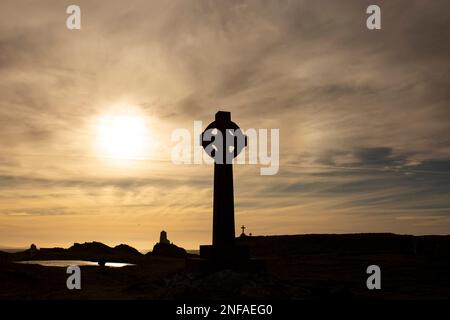 Silhouette d'une croix celtique avec le soleil du soir directement derrière elle. Avec un phare, des bâtiments et une croix en arrière-plan Banque D'Images