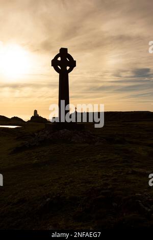 Silhouette d'une croix celtique avec le soleil du soir directement derrière elle. Avec un phare, des bâtiments et une croix en arrière-plan Banque D'Images