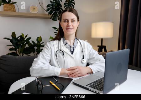 Portrait d'une jeune femme brune souriante et mignonne assise à un bureau à l'hôpital ou dans une clinique privée. Bonne positive caucasienne femme médecin dans blanc med Banque D'Images