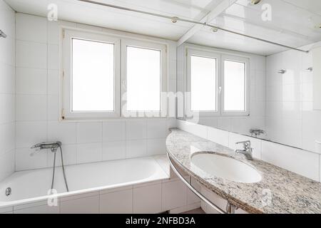 Salle de bains blanche avec grand bain à remous. Sur le mur se trouvent deux fenêtres en plastique avec verre dépoli à travers lequel la lumière du jour brille. Tout près, il y a un lavabo sur marbre Banque D'Images