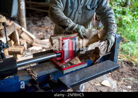 Un homme de race blanche coupe le bois avec un séparateur de rondins hydraulique dans une chemise en denim, des gants de travail et des protections auditives. Velours de bois défoqué derrière Banque D'Images