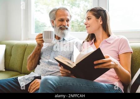 Un homme âgé souriant aime passer son temps avec sa petite-fille, une jeune femme assise à côté de lui tenant un livre Banque D'Images