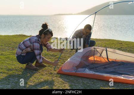 Couple asiatique préparant une tente pour camper dans la pelouse avec le lac en arrière-plan pendant le coucher du soleil Banque D'Images