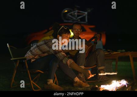 Femme asiatique en train de griller des guimauves tandis que son petit ami joue de la guitare au feu de camp où ils ont installé leurs tentes pour camper au bord du lac la nuit. Banque D'Images