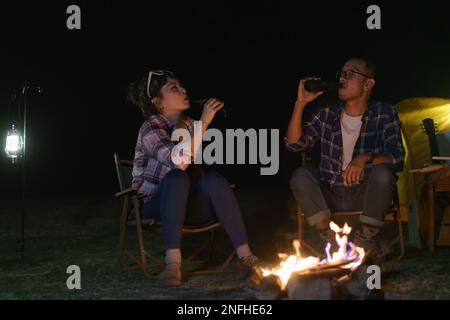 Un couple asiatique buvant de la bière avec un feu de camp, ils se sont mis en place pour éclairer la nuit au milieu d'une tente de camping près d'un lac. Banque D'Images