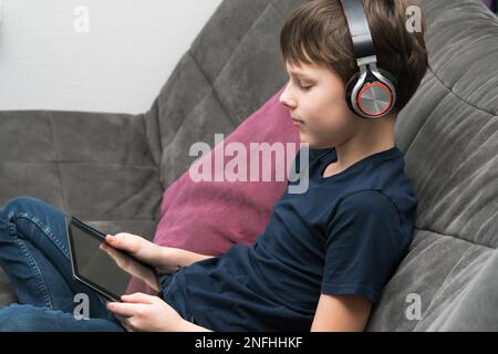 Joyeux et spirituel petit garçon dans le casque regarder des films et écouter de la musique avec la tablette numérique et le service de streaming, asseyez-vous sur le canapé gris. Livres audio Banque D'Images