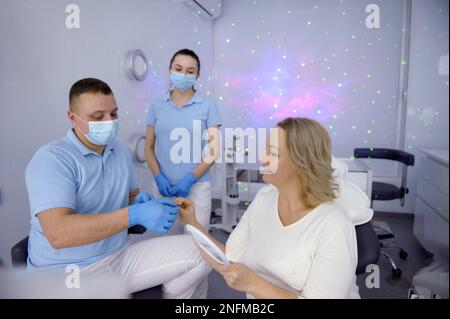 clinique dentaire réception des patients dans le hall avec des étoiles atmosphère intime médecin et infirmière dans les masques portant des gants bleus offrent aux femmes options de couleur blanchiment des dents technologie la plus récente Banque D'Images