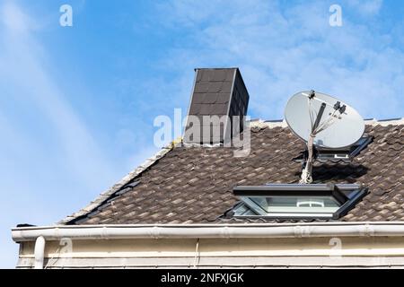 Un fragment d'un toit en tuiles d'une maison avec cheminée, parabole satellite et fenêtre contre un ciel bleu avec des nuages de lumière. Gros plan. Banque D'Images