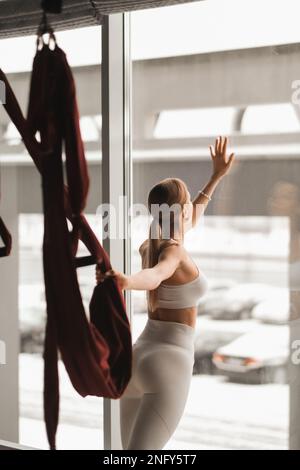 Portrait d'une fille en vêtements de sport blancs debout près de la fenêtre et tenant un hamac suspendu dans la main dans la salle de fitness. Banque D'Images