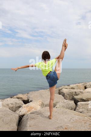 Jeune fille adolescente mince faisant des exercices sur les rochers pour garder la forme en été près de la mer avec sa jambe relevée Banque D'Images