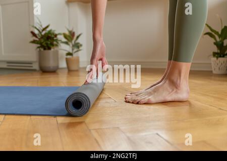 Vue latérale d'une femelle pieds nus non reconnaissable dans des leggings kaki de sport qui se déroulent sur un tapis de yoga bleu, debout sur un plancher en bois, pour préparer une séance de yoga. Banque D'Images