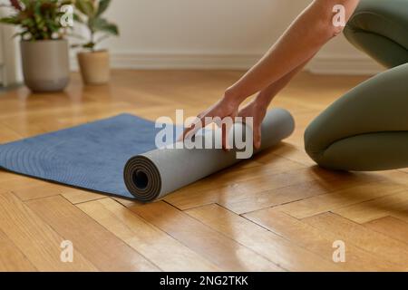 Photo courte d'une femme anonyme en train de dérouler un tapis de yoga bleu pour se préparer à une séance de yoga sur un plancher de bois franc dans le salon à l'intérieur. Banque D'Images