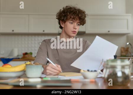 Gros plan d'un jeune homme aux cheveux bouclés portant un t-shirt beige assis sur une table de cuisine entourée de confort de la vie domestique pour prendre des notes. Banque D'Images