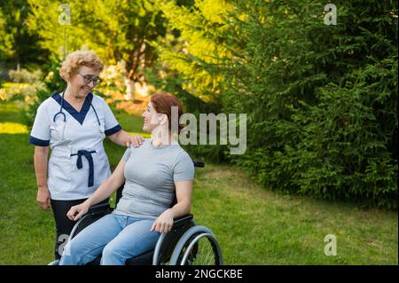 Une infirmière âgée marche avec une femme d'âge moyen en fauteuil roulant dans le parc. Banque D'Images