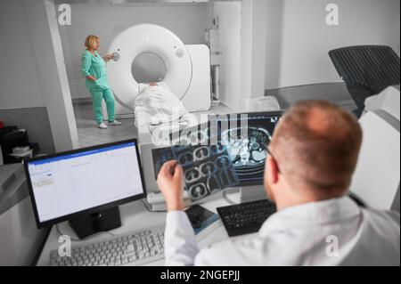 Scanner médical de tomodensitométrie ou IRM. Vue arrière d'un médecin de sexe masculin assis à l'ordinateur, infirmière faisant l'IRM. Concentrez-vous sur l'infirmière. Spécialistes travaillant à l'hôpital, étudiant les résultats, examinant. Banque D'Images