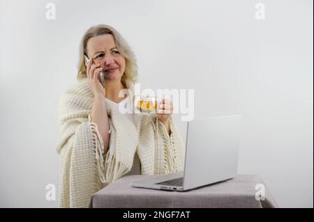 Happy Millenium gen Z smartphone utilisateur fille enveloppée dans un tissu écossais chaud, boire du thé chaud, café, cacao, en utilisant l'application en ligne sur smartphone, se détendre sur le canapé dans la maison confortable. Photo de haute qualité Banque D'Images