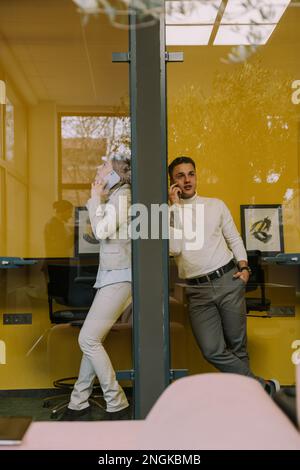 Deux collègues masculins et féminins attrayants ayant un appel téléphonique dans les cabines téléphoniques au bureau Banque D'Images