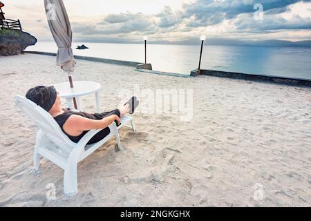 Détendez-vous dans une chaise longue, à côté d'une plage tropicale, en regardant le soleil doré se coucher derrière des nuages qui s'ensuivent, à travers la mer des Philippines, se sentant calme et p Banque D'Images