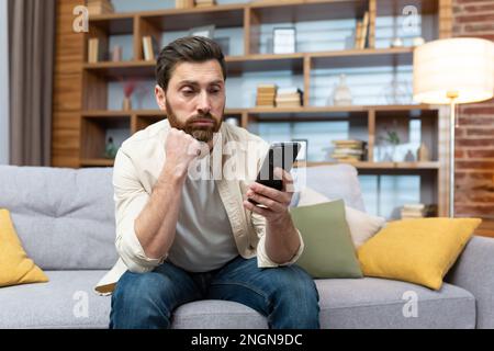 Triste et déprimé homme seul à la maison assis sur un canapé, indépendant dans des vêtements décontractés tenant un smartphone, sans satisfaction lisant les mauvaises nouvelles en ligne à partir du téléphone. Banque D'Images