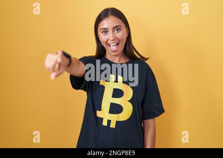 Jeune femme hispanique portant un t-shirt bitcoin pointant vers vous et l'appareil photo avec les doigts, souriant positif et gai Banque D'Images